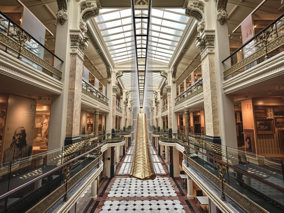 This is a picture taken from the mezzanine level of the Luce Foundation Center for American Art with an artwork by Glenn Kaino centered. There are cases filled with sculptures and paintings on the right and left side of the image, and a skylight illuminating the space.