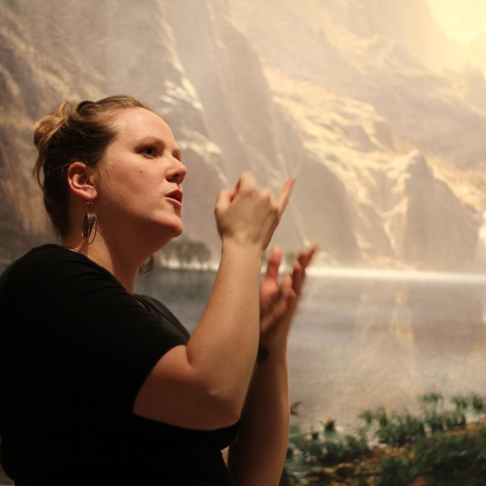 A photograph of a woman using her hands to sign in front of a landscape painting.