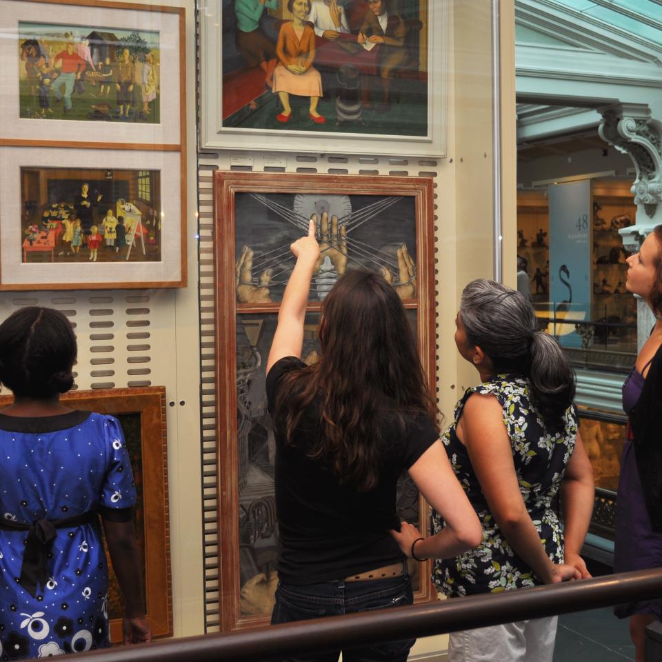 Four museum visitors stand in front of a case full of paintings in the Luce Foundation Center. One visitor raises their hand to point at a painting.