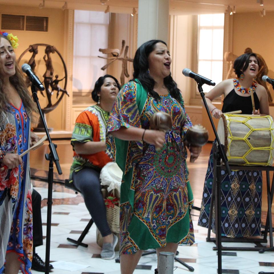 This is an image of the band La Marvela. Four band members dressed in colorful attire sing into microphones and play instruments.