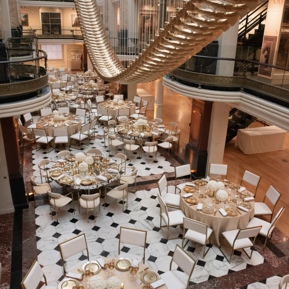 This is an image of the Luce Foundation Center set up for a special event. Tables, dishware, and chairs are spread out across the black and white marble flooring and an artwork with gilded arms hangs overhead.