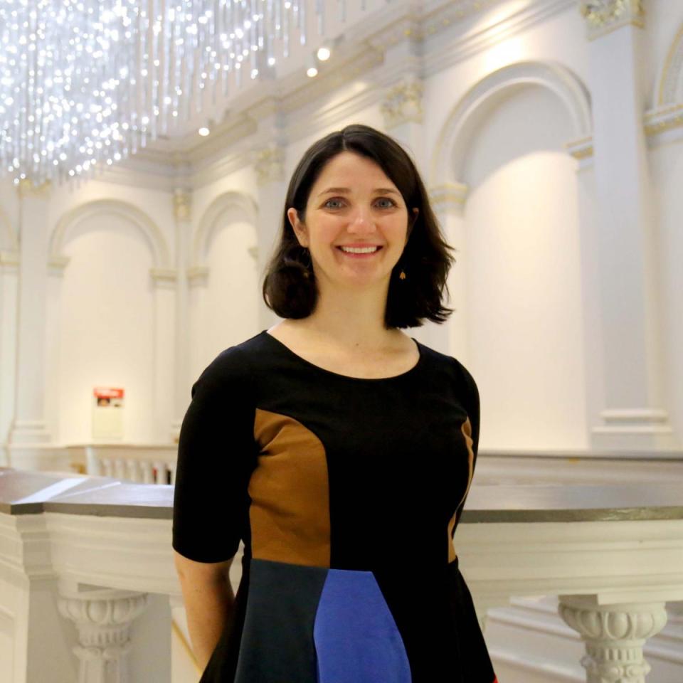 A photograph of a woman with brown hair and a dress standing inside a building.