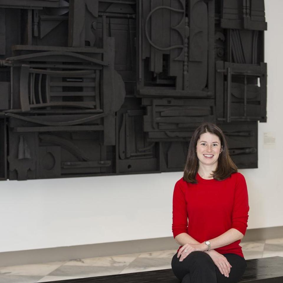 A photograph of a woman sitting down on a bench in a gallery with a red sweater on and a black artwork behind her.