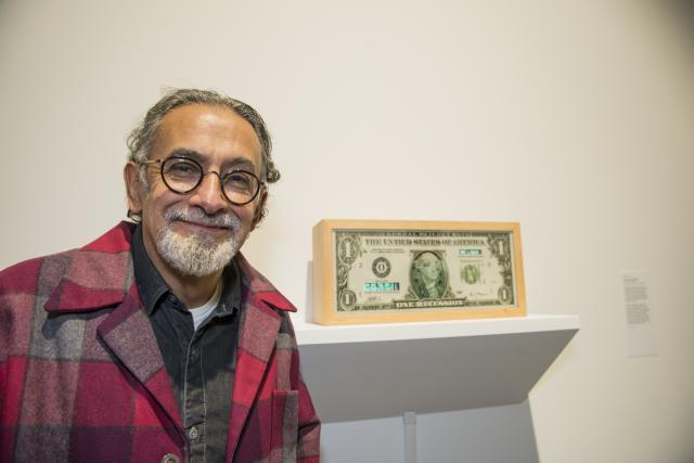 A man with a white beard standing in front of a framed one dollar bill