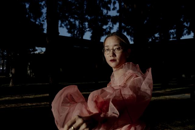 Christine Sun Kim Portrait Christine Sun Kim stands in front of a tree and dark background in a pink dress