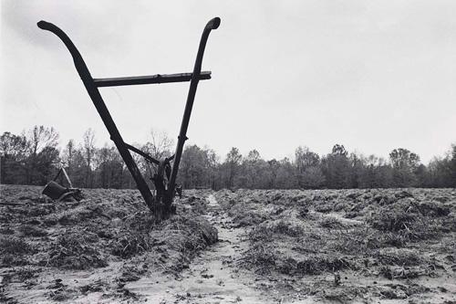 An gelatin silver print of a muddy field with a farming tool.