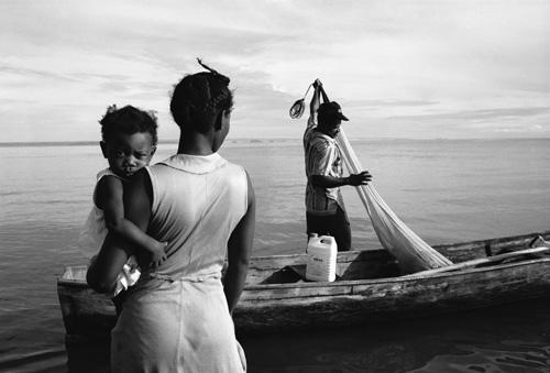 Gleaton's gelatin silver print of a woman holding a baby while watching a man in a fishing boat.
