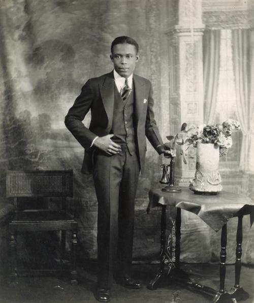 VanDerZee's gelatin silver print of a man standing next to a table with flowers and a telephone.