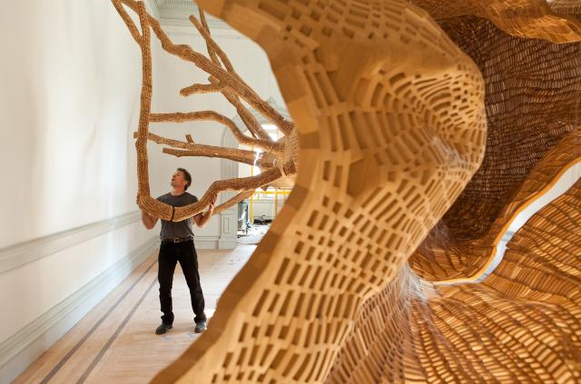 A photograph of a man holding a limb of a tree inside of a room.