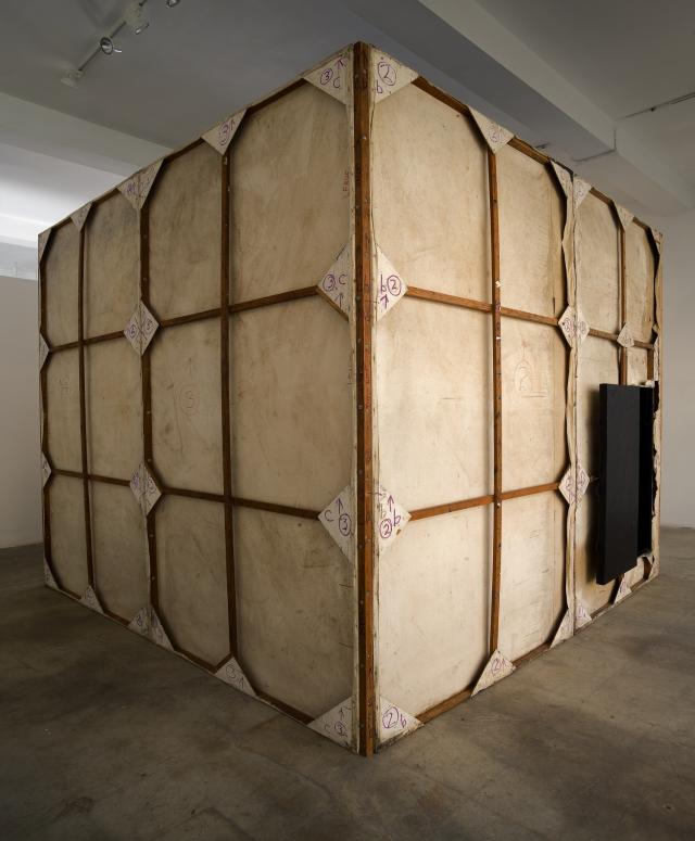 Exterior photograph of a tan, cube-shaped structure with a black entrance door