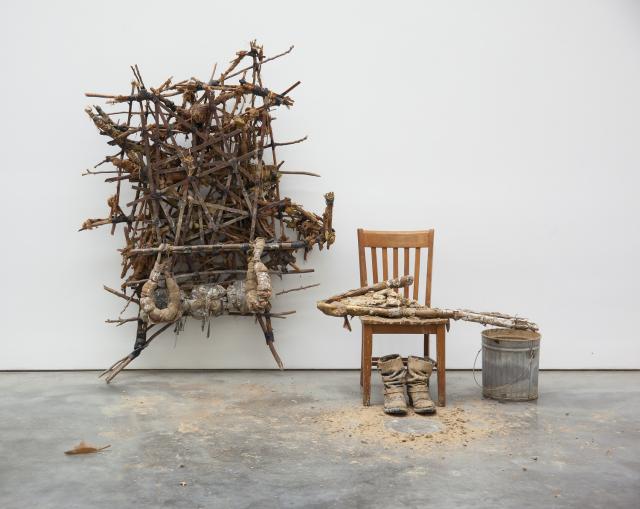Sculpture formed from sticks, mud, and rope on a wall, with a chair and bucket positioned to the right of it 
