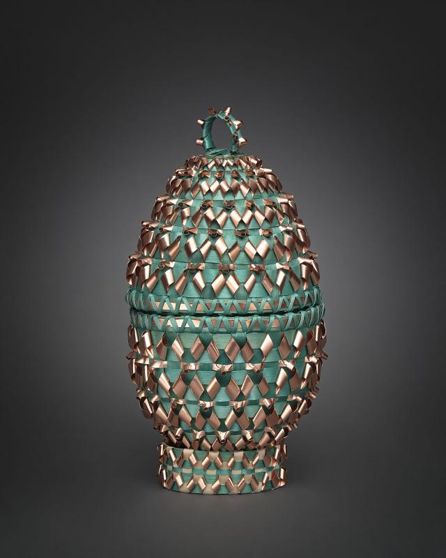 A oval artwork with green and gold material woven into it. 