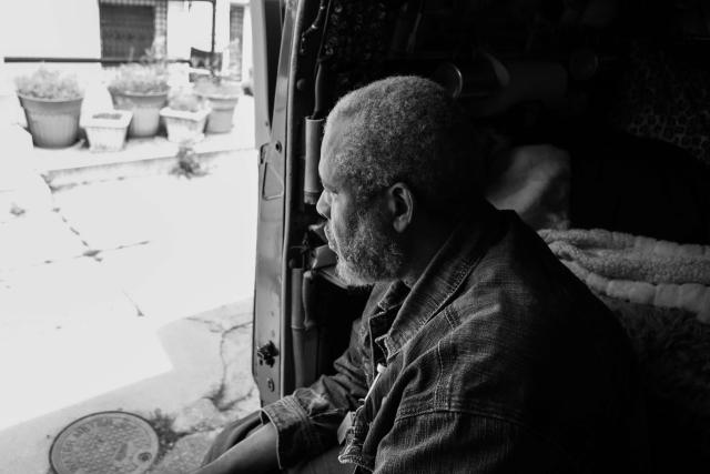 Black and white photo of an African American man sitting in the shade of a car interior, looking out onto a brightly lit sidewalk.