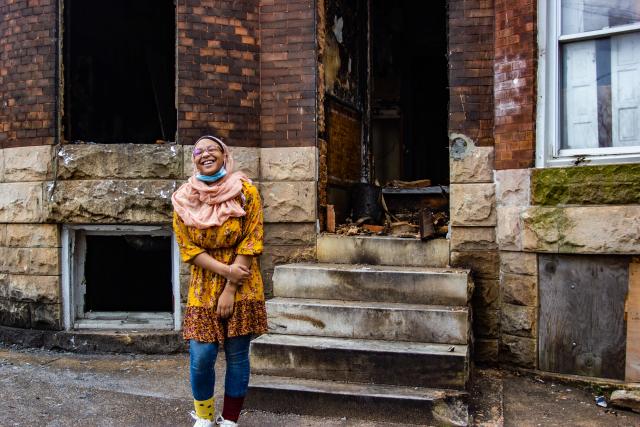 An African American girl wearing a rose-colored hijab stands, smiling, in front of a stone and brick rowhome.