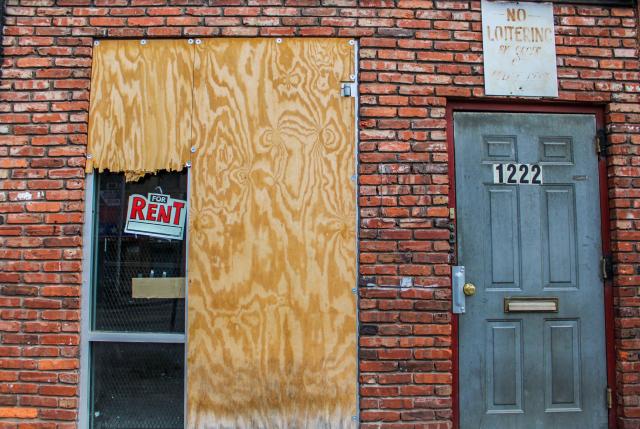 The exterior of a building with two doors, one boarded up with a "For Rent" sign to the left and one with a "No Loitering" sign above.