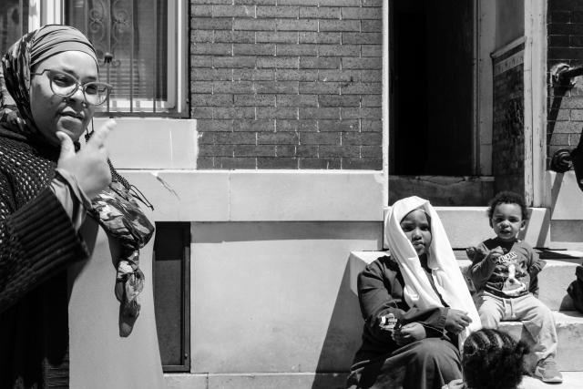 Four people outside a brick rowhome. A woman and girl wearing hijabs as well as a young child look directly at the camera. The back of a child's braided hair can be seen. 