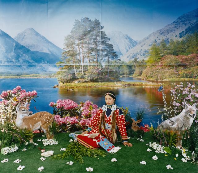 Native woman sitting on a grassy patch with a blow up deer next to her and a mountain landscape