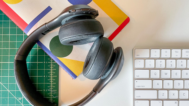 The surfce of a desk with headphones, keyboard, notebook, and cutting board.
