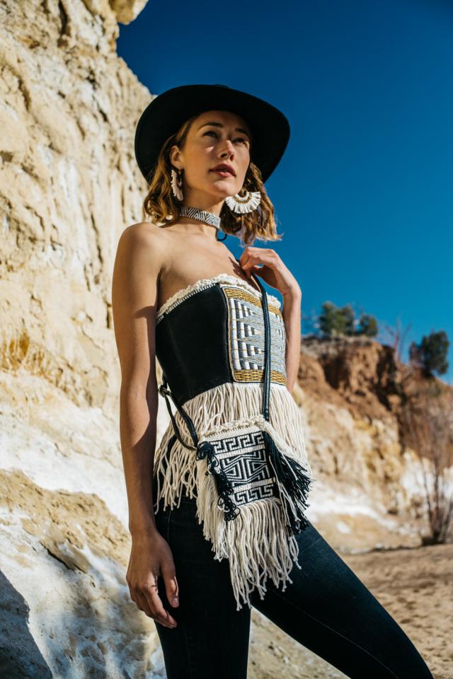 A woman stands in front of a rocky desert landscape. She's wearing a black and white hand-woven ensemble.