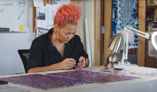 Artist Erica Lord sits at a table. She is beading.