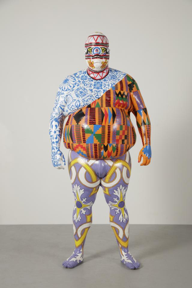 Sculpture of a person completely covered with multiple colorful, intricate patterns standing against a neutral background.