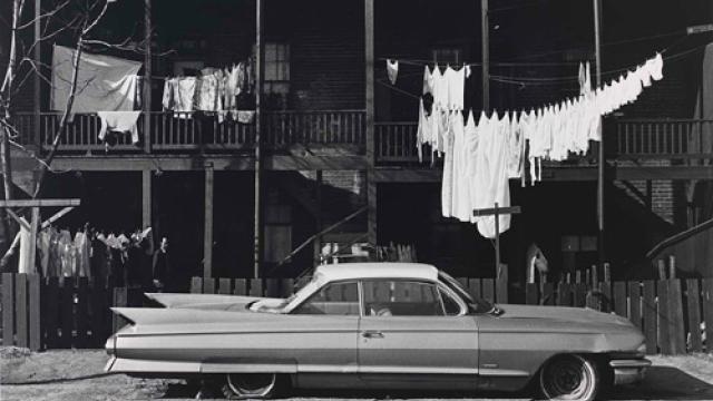 A gelatin silver print of a street with a car in the foreground, clothes handing in the middle ground and a building in the background.