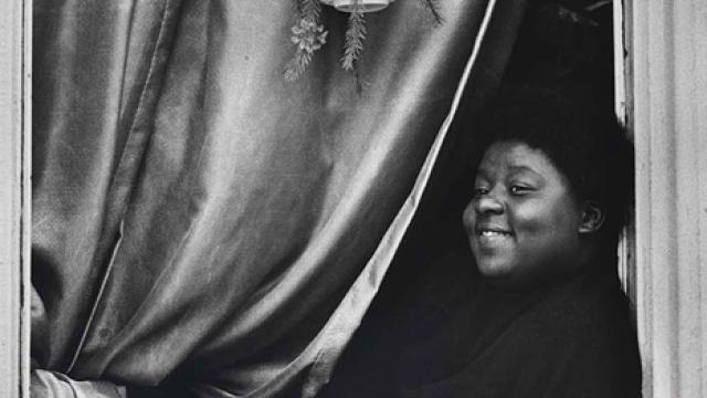 A gelatin silver print of a woman leaning in her window looking out with a curtain behind her. 