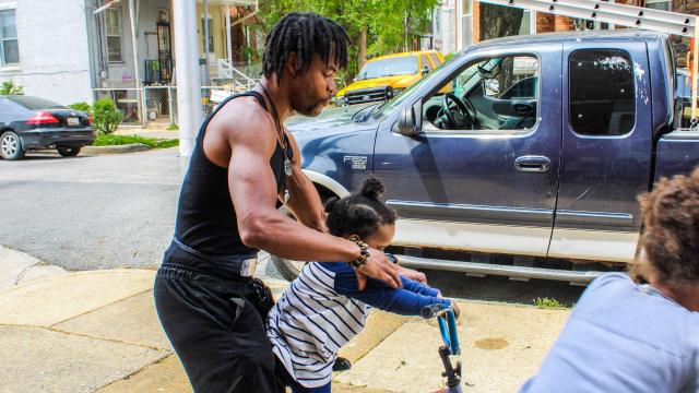 A muscular African American man helps a small child ride a large bike. Another child can be seen at the extreme foreground. Work trucks are parked along the street.