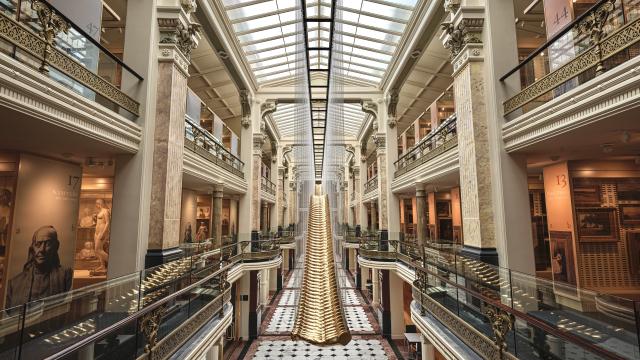 This is a picture taken from the mezzanine level of the Luce Foundation Center for American Art with an artwork by Glenn Kaino centered. There are cases filled with sculptures and paintings on the right and left side of the image, and a skylight illuminating the space.