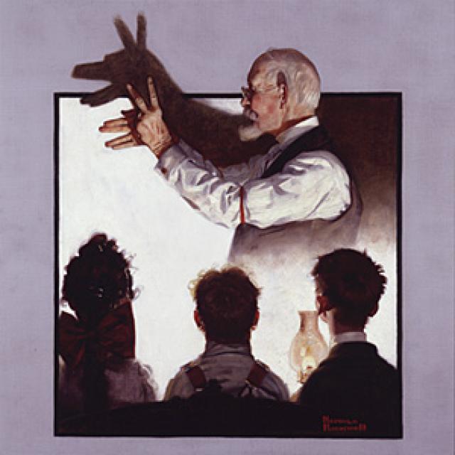 An image of a man making shadow puppets on the wall in the background and three children watching in the foreground. 