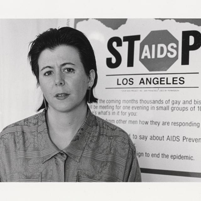 A black and white photograph of a person with short hair standing in front of a sign that says "Stop AIDS Los Angeles."