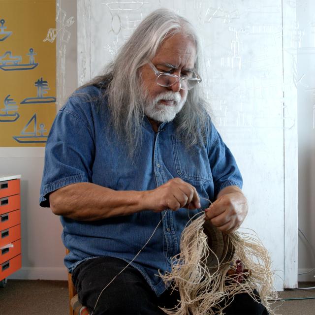 A man with long white hair and a beard sits and works on a straw like material.
