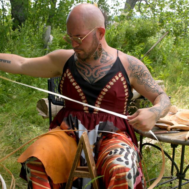 A bald man whose arms are covered in tattoos sits outside. He is holding canes for basketry.