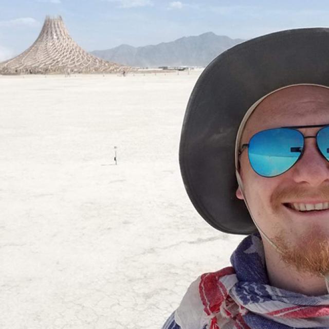 A man wearing a hat, sunglasses, and a scarf taking a selfie in the desert. A pyre is in the distance.