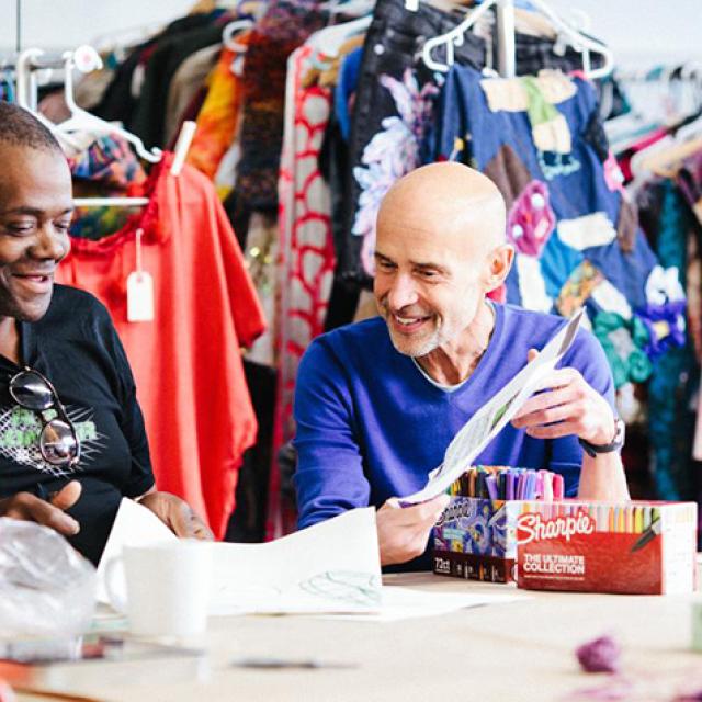 Three people sit at a table. Racks of clothes hang behind them.