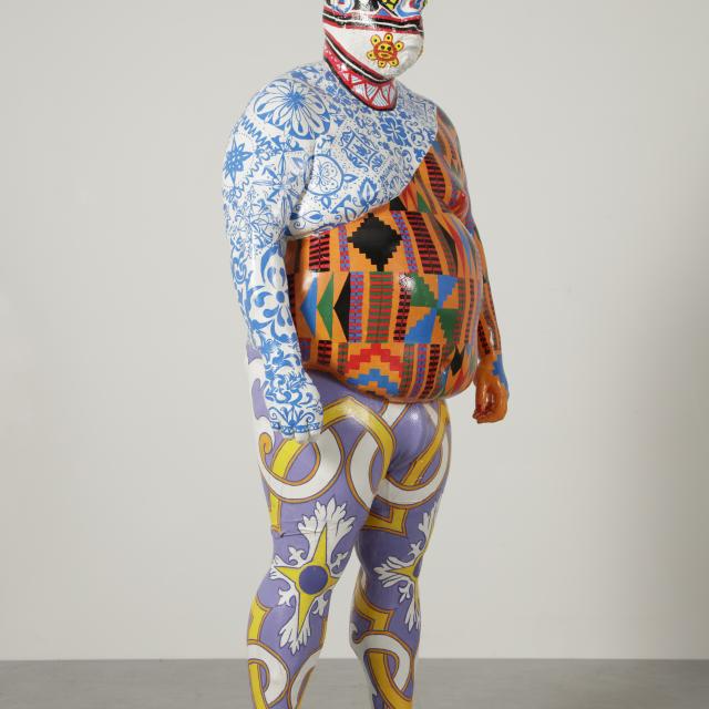 Sculpture of a person completely covered with multiple colorful, intricate patterns standing against a neutral background.