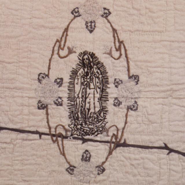 Embroidered tapestry with the Virgen de Guadalupe in the center, flowers around the frame, and barbed wire slashing through the middle of the work.