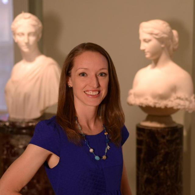 A photograph of a woman standing in an art gallery with busts behind her. 