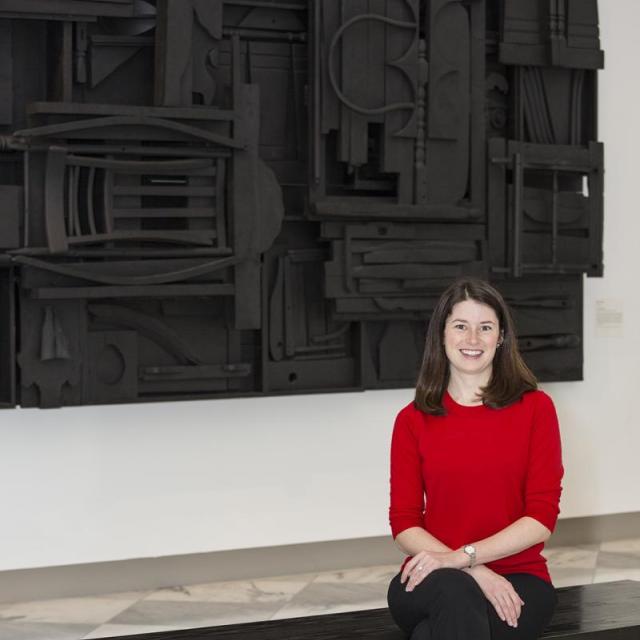 A photograph of a woman sitting down on a bench in a gallery with a red sweater on and a black artwork behind her.